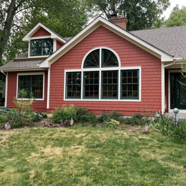 A beautifully painted red shingle home with white trim and green-framed windows, set in a lush yard with small American flags, showcasing the curb appeal achieved through expert exterior painting.