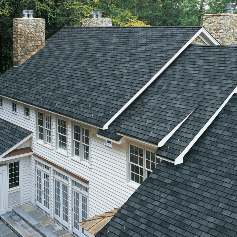 An aerial view of a clean, dark gray asphalt shingle roof on a white-sided home with stone chimneys, showcasing the results of expert roof cleaning and preservation.