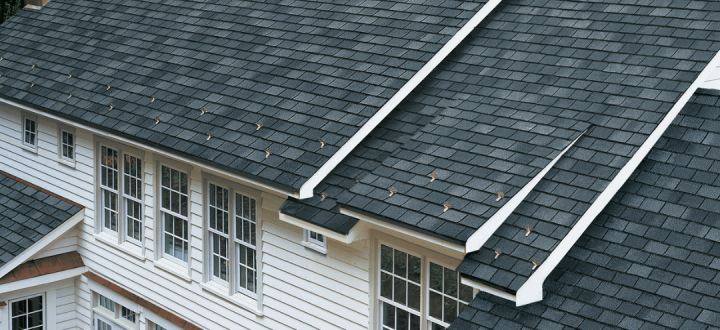 Aerial view of a clean, dark gray asphalt shingle roof on a white-sided home with stone chimneys, showcasing the results of professional roof pressure washing and maintenance.