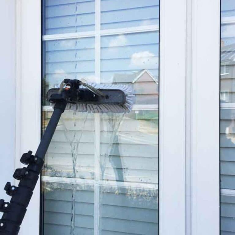 A water-fed pole with a brush attachment cleans a large window on a residential home, demonstrating a safe and effective method for exterior window washing without ladders or scaffolding.