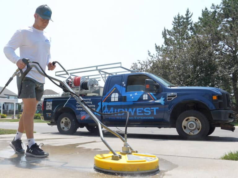 A technician from Midwest Prowash and Exteriors and Exteriors uses a surface cleaning machine on a driveway, with the company's branded blue work truck in the background.